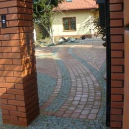 Tomasbruk USLUGI BRUKARSKIE USŁUGI REMONTOW-BUDOWLANE - Ułożona z kostki brukowej, zakrzywiona ścieżka prowadząca do domu, widoczna przez ceglany filar bramy wejściowej. Użyto czerwonej i szarej kostki, tworząc dekoracyjny wzór.